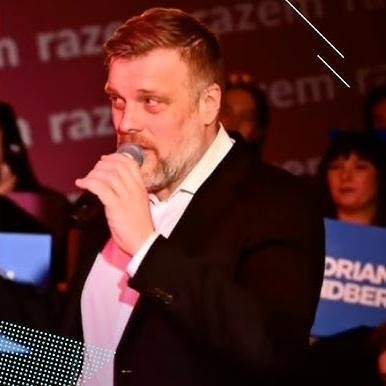 Adrian Zandberg w zamyśleniu, w otoczeniu sugerującym jego działalność polityczną.