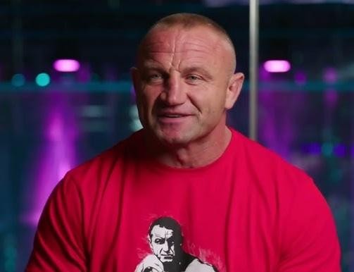 Mariusz Pudzianowski w dynamicznej pozie podczas walki MMA lub treningu siłowego, ukazujący jego muskularną sylwetkę i determinację.